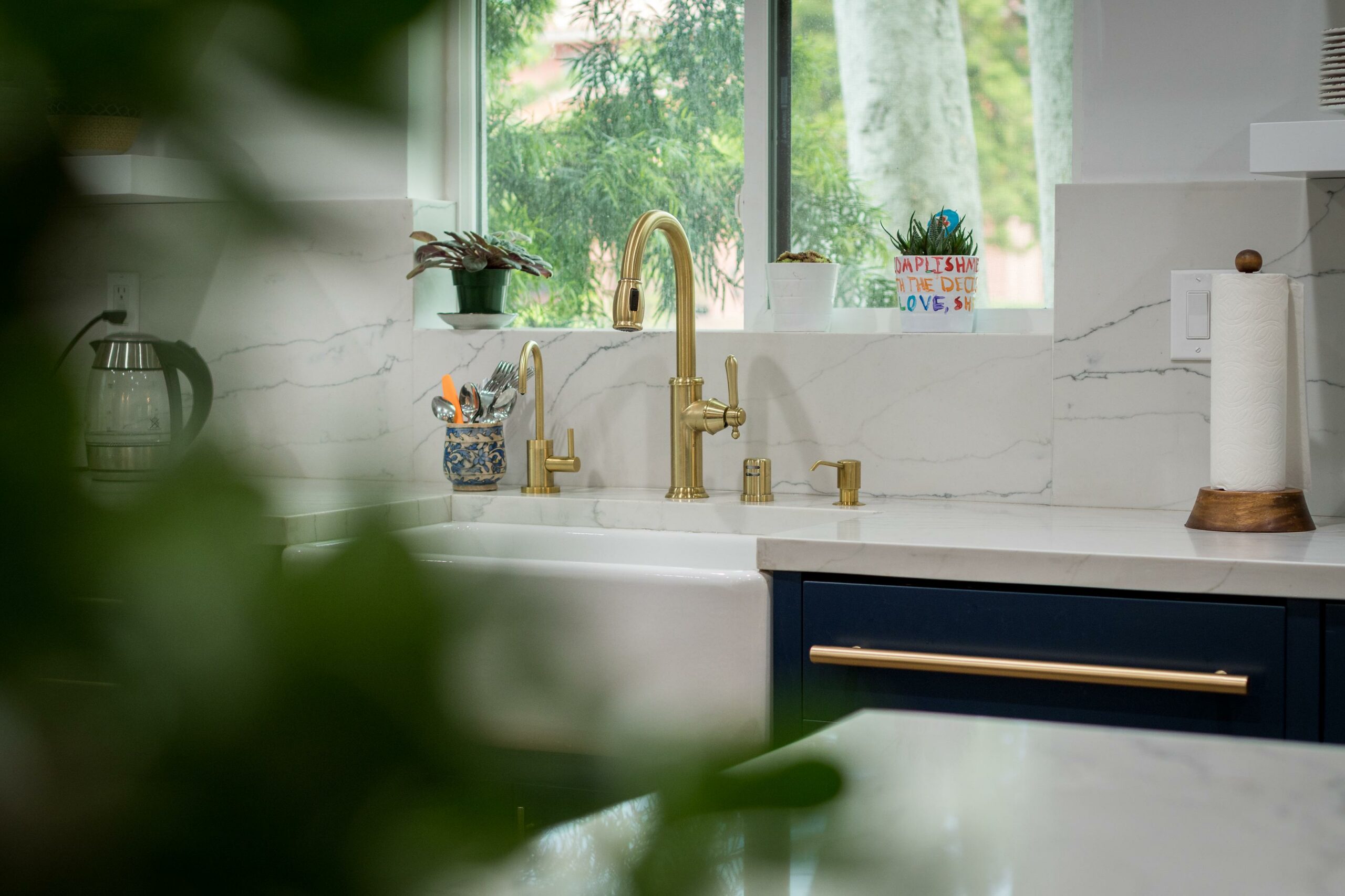 Modern kitchen sink with gold faucet and greenery outside.